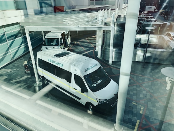 Two ambulances are parked outside what appears to be a modern building. The view is from above and through a window, creating reflections and a slightly obstructed view. The ground is paved with patterned tiles and there is a partially visible shelter above the ambulances. The scene appears calm with no immediate activity around the vehicles.
