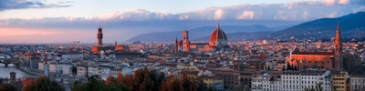 A panoramic view of an Italian cityscape during sunset, featuring prominent historical architecture including a large dome, tall tower, and a river running alongside the buildings. The architecture includes a mix of red rooftops, beige walls, and ornate structures. Soft clouds and distant hills are visible in the background, creating a harmonious blend of natural and urban elements.