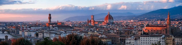 A panoramic view of an Italian cityscape during sunset, featuring prominent historical architecture including a large dome, tall tower, and a river running alongside the buildings. The architecture includes a mix of red rooftops, beige walls, and ornate structures. Soft clouds and distant hills are visible in the background, creating a harmonious blend of natural and urban elements.