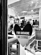 A black heart-shaped sticker is displayed on a window, with the words 'FOR UKRAINE WE STAND' in bold white letters. The interior of a shop or gallery is visible through the glass, with various items and artworks displayed. An American Express sticker is also seen in the bottom left corner of the window.