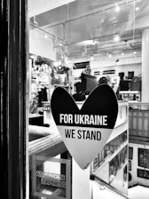 A black heart-shaped sticker is displayed on a window, with the words 'FOR UKRAINE WE STAND' in bold white letters. The interior of a shop or gallery is visible through the glass, with various items and artworks displayed. An American Express sticker is also seen in the bottom left corner of the window.