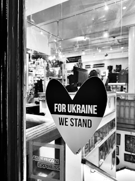 A black heart-shaped sticker is displayed on a window, with the words 'FOR UKRAINE WE STAND' in bold white letters. The interior of a shop or gallery is visible through the glass, with various items and artworks displayed. An American Express sticker is also seen in the bottom left corner of the window.