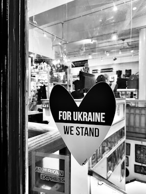 A black heart-shaped sticker is displayed on a window, with the words 'FOR UKRAINE WE STAND' in bold white letters. The interior of a shop or gallery is visible through the glass, with various items and artworks displayed. An American Express sticker is also seen in the bottom left corner of the window.