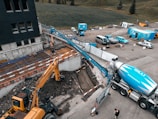A construction site featuring a large cement truck connected to a pump with a long blue hose, extending to a construction area where workers are present. A yellow excavator is also situated nearby, surrounded by construction equipment and materials. The site is adjacent to a field and a multi-story building. Several vehicles and storage containers are visible in the background.