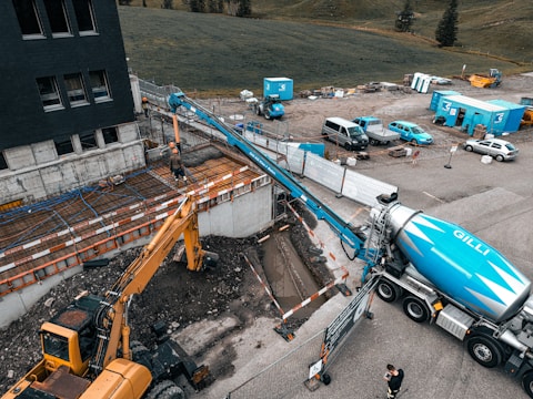 A construction site featuring a large cement truck connected to a pump with a long blue hose, extending to a construction area where workers are present. A yellow excavator is also situated nearby, surrounded by construction equipment and materials. The site is adjacent to a field and a multi-story building. Several vehicles and storage containers are visible in the background.