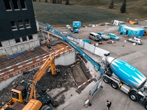 A construction site featuring a large cement truck connected to a pump with a long blue hose, extending to a construction area where workers are present. A yellow excavator is also situated nearby, surrounded by construction equipment and materials. The site is adjacent to a field and a multi-story building. Several vehicles and storage containers are visible in the background.