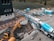 A construction site featuring a large cement truck connected to a pump with a long blue hose, extending to a construction area where workers are present. A yellow excavator is also situated nearby, surrounded by construction equipment and materials. The site is adjacent to a field and a multi-story building. Several vehicles and storage containers are visible in the background.