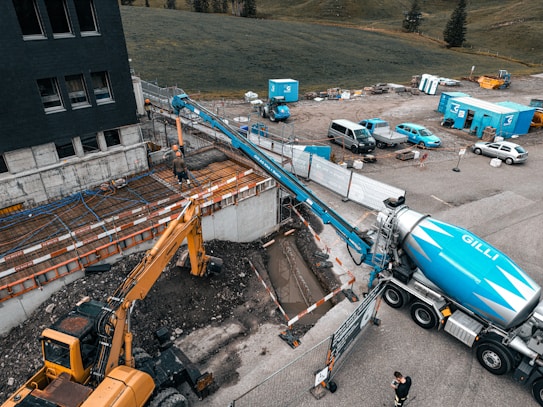 A construction site featuring a large cement truck connected to a pump with a long blue hose, extending to a construction area where workers are present. A yellow excavator is also situated nearby, surrounded by construction equipment and materials. The site is adjacent to a field and a multi-story building. Several vehicles and storage containers are visible in the background.