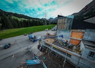 A construction site near a road with vehicles and machinery in operation. Workers are present, using equipment to build or renovate a structure. The site is surrounded by a rural landscape with grass, trees, and mountains under a partly cloudy sky.