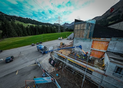 A construction site near a road with vehicles and machinery in operation. Workers are present, using equipment to build or renovate a structure. The site is surrounded by a rural landscape with grass, trees, and mountains under a partly cloudy sky.