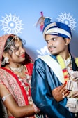 A couple dressed in traditional Indian wedding attire, with the woman wearing a red and gold saree adorned with intricate embroidery and jewelry, while the man is in a blue and white sherwani with a decorative turban. They are exchanging a joyful and affectionate look, captured against a light blue background with ornamental patterns.