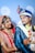 A couple dressed in traditional Indian wedding attire, with the woman wearing a red and gold saree adorned with intricate embroidery and jewelry, while the man is in a blue and white sherwani with a decorative turban. They are exchanging a joyful and affectionate look, captured against a light blue background with ornamental patterns.