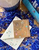 A decorative setup featuring blue shredded paper and colorful star confetti scattered around. Two blue colored books with intricate designs and titles are placed on the right. A cardboard envelope with a mandala pattern and a silver necklace with a state-shaped pendant lies on top of a white envelope. A decorative candle, partially in view, is to the left.