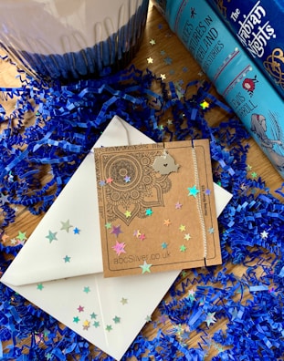 A decorative setup featuring blue shredded paper and colorful star confetti scattered around. Two blue colored books with intricate designs and titles are placed on the right. A cardboard envelope with a mandala pattern and a silver necklace with a state-shaped pendant lies on top of a white envelope. A decorative candle, partially in view, is to the left.