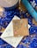A decorative setup featuring blue shredded paper and colorful star confetti scattered around. Two blue colored books with intricate designs and titles are placed on the right. A cardboard envelope with a mandala pattern and a silver necklace with a state-shaped pendant lies on top of a white envelope. A decorative candle, partially in view, is to the left.