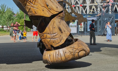 A large, intricately carved wooden robotic structure takes center stage in an outdoor urban setting. Several people, including adults and children, are observing and interacting with the scene. The background features industrial elements and a cylindrical building.