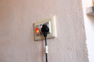 An electrical wall socket with a black power plug inserted. The switch next to the socket is turned on, indicated by a red light. The wall has a textured surface with a light beige color.