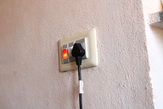 An electrical wall socket with a black power plug inserted. The switch next to the socket is turned on, indicated by a red light. The wall has a textured surface with a light beige color.