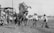 A dynamic scene featuring a rearing horse, surrounded by several people, two of whom are attempting to handle the animal. Onlookers are present in the background, standing near a wooden structure. The setting appears to be an outdoor event, possibly a rodeo or similar show.