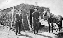 Two men stand in front of a sod house, both wearing hats and work clothes. One man holds a tool, possibly a hammer. A horse is harnessed to the right, suggesting a rural, agrarian setting. There are various tools and remnants of construction around the area.