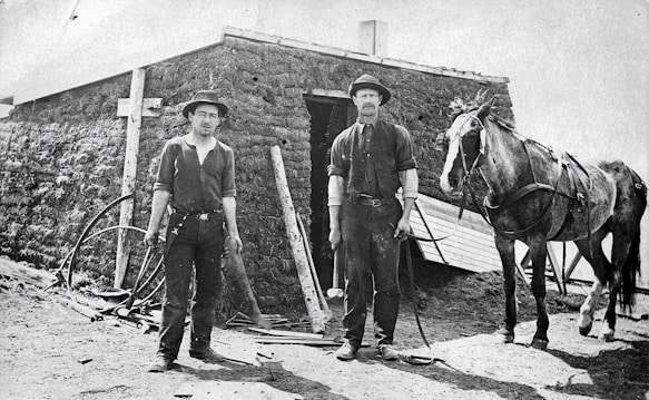 Two men stand in front of a sod house, both wearing hats and work clothes. One man holds a tool, possibly a hammer. A horse is harnessed to the right, suggesting a rural, agrarian setting. There are various tools and remnants of construction around the area.