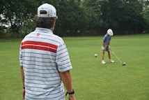 A person wearing a striped polo shirt and a cap stands on a golf course, observing another individual in a helmet preparing to hit a golf ball. The scene is set outdoors on a lush, green lawn surrounded by trees.