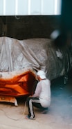 A person wearing a white cap and a white shirt is crouching near the rear end of a car in a garage, applying paint with a spray gun. The car is partially covered with protective plastic sheets and masking paper.