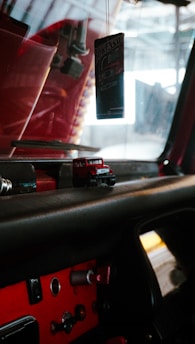 The image depicts the interior of a vehicle, focusing on the dashboard area. There is a small toy truck on the dashboard, and an air freshener hanging from the rearview mirror with a design that includes the words 'RokenrSoll'. The dashboard is mainly black with some red accents and has various buttons and controls.