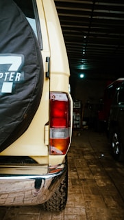 The image presents the rear view of a beige off-road vehicle inside a dimly lit garage. A spare tire with a black cover featuring white lettering is mounted on the back door. The vehicle's rear light is visible, and the ground is covered with brick paving. Another vehicle is partially visible to the right.