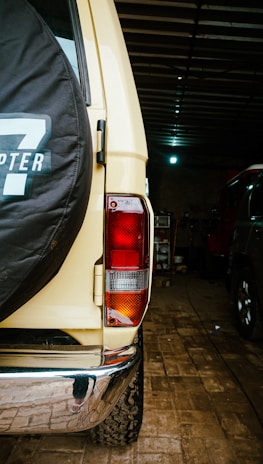 The image presents the rear view of a beige off-road vehicle inside a dimly lit garage. A spare tire with a black cover featuring white lettering is mounted on the back door. The vehicle's rear light is visible, and the ground is covered with brick paving. Another vehicle is partially visible to the right.