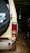 The image presents the rear view of a beige off-road vehicle inside a dimly lit garage. A spare tire with a black cover featuring white lettering is mounted on the back door. The vehicle's rear light is visible, and the ground is covered with brick paving. Another vehicle is partially visible to the right.
