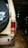 The image presents the rear view of a beige off-road vehicle inside a dimly lit garage. A spare tire with a black cover featuring white lettering is mounted on the back door. The vehicle's rear light is visible, and the ground is covered with brick paving. Another vehicle is partially visible to the right.