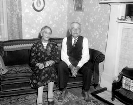 An elderly couple sits together on a plush, patterned couch in a room with floral wallpaper. The woman wears a dark floral dress and holds her hands in her lap, while the man, dressed in a vest and tie, has his hands clasped. A small fireplace and decorative items are visible in the background, adding a cozy, homely atmosphere.
