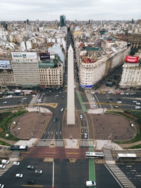 A large cityscape view featuring a tall obelisk at the center of a busy intersection, surrounded by high-rise buildings and numerous advertisements. The street layout includes several lanes with vehicles and pedestrian paths. The urban environment is expansive, and the sky is overcast, suggesting a typical day in a bustling city.