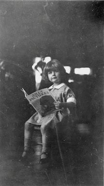 A young child is seated on a stool, holding a book titled 'Jolly Pictures'. The child is dressed in a light-colored dress with a white collar and has a calm and curious expression. The background is dark, with highlights that suggest a softly lit environment.