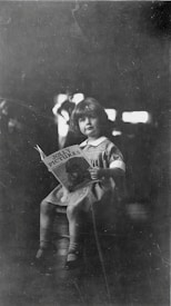 A young child is seated on a stool, holding a book titled 'Jolly Pictures'. The child is dressed in a light-colored dress with a white collar and has a calm and curious expression. The background is dark, with highlights that suggest a softly lit environment.