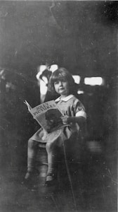 A young child is seated on a stool, holding a book titled 'Jolly Pictures'. The child is dressed in a light-colored dress with a white collar and has a calm and curious expression. The background is dark, with highlights that suggest a softly lit environment.