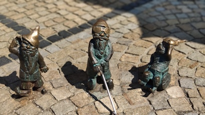 Three small bronze statues are arranged on a cobblestone paving. The figures appear to be gnomes or dwarfs, each with unique attributes: one holds a hat, another uses a walking stick, and the third is seated with a small creature on its head.