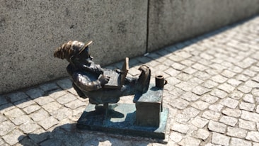 A small bronze sculpture of a gnome reclining on a chair with its legs up on a table, using a laptop. It is placed on a cobblestone surface and is illuminated by bright sunlight. A small cup or mug is also situated on the table beside the gnome.