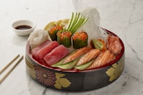 A beautifully arranged platter of sashimi featuring various types of raw fish, including salmon and tuna, topped with garnish and accompanied by roe. The dish is presented in a decorative bowl with floral patterns. Thin slices of cucumber, lemon, and ginger are also included, alongside a small dish of soy sauce. Chopsticks are placed on the side on a white marble surface.