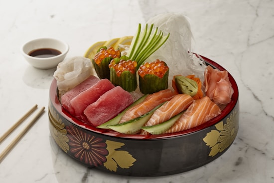 A beautifully arranged platter of sashimi featuring various types of raw fish, including salmon and tuna, topped with garnish and accompanied by roe. The dish is presented in a decorative bowl with floral patterns. Thin slices of cucumber, lemon, and ginger are also included, alongside a small dish of soy sauce. Chopsticks are placed on the side on a white marble surface.