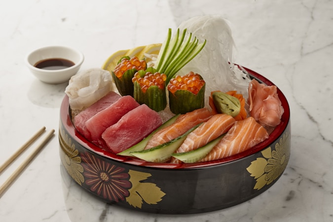 A beautifully arranged platter of sashimi featuring various types of raw fish, including salmon and tuna, topped with garnish and accompanied by roe. The dish is presented in a decorative bowl with floral patterns. Thin slices of cucumber, lemon, and ginger are also included, alongside a small dish of soy sauce. Chopsticks are placed on the side on a white marble surface.