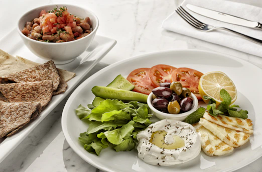 A white plate with a Mediterranean assortment including sliced cucumbers, tomatoes, a lemon wedge, black and green olives, mint leaves, grilled halloumi slices, and a dollop of thick yogurt with a sprinkle of herbs and olive oil. Next to it, there is a smaller plate with toasted flatbread. A bowl filled with mixed beans and diced tomatoes sits close by, accompanied by a set of cutlery on a napkin.