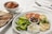 A white plate with a Mediterranean assortment including sliced cucumbers, tomatoes, a lemon wedge, black and green olives, mint leaves, grilled halloumi slices, and a dollop of thick yogurt with a sprinkle of herbs and olive oil. Next to it, there is a smaller plate with toasted flatbread. A bowl filled with mixed beans and diced tomatoes sits close by, accompanied by a set of cutlery on a napkin.