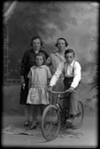 A vintage black and white photograph featuring two women standing in the background, one with her hand on the other's shoulder, and two children in the foreground. One child, a boy, is sitting on a bicycle with a determined expression, while the other child, a girl, stands beside him wearing a dress. The background is painted to resemble an outdoor scene with foliage.