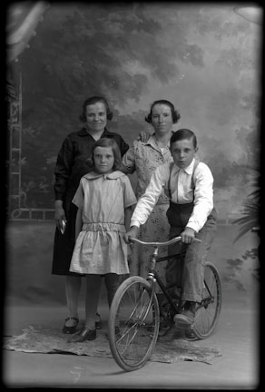 A vintage black and white photograph featuring two women standing in the background, one with her hand on the other's shoulder, and two children in the foreground. One child, a boy, is sitting on a bicycle with a determined expression, while the other child, a girl, stands beside him wearing a dress. The background is painted to resemble an outdoor scene with foliage.