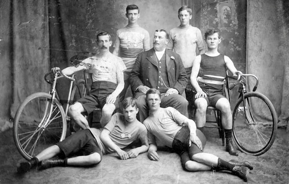A vintage black-and-white photograph featuring seven men in athletic attire, arranged in a studio setting with a patterned backdrop. Two of the men are holding bicycles, suggesting a theme related to cycling. The central figure, seated in a suit, appears to be of higher status or a manager. The overall attire and style indicate the early 20th century.