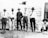 Six men are standing outside a rustic, wooden building in a rural setting. Most are wearing broad-brimmed hats, buttoned shirts, and long pants, with one man wearing woolly chaps. The scene has an old-fashioned, historical feel to it, with the men posing casually.