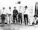 Six men are standing outside a rustic, wooden building in a rural setting. Most are wearing broad-brimmed hats, buttoned shirts, and long pants, with one man wearing woolly chaps. The scene has an old-fashioned, historical feel to it, with the men posing casually.