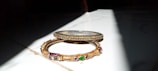 Two ornate bangles are placed on a surface partially illuminated by sunlight. One bangle is slim with green stones embedded around it, while the other is wider with intricate gold designs and multi-colored stones.